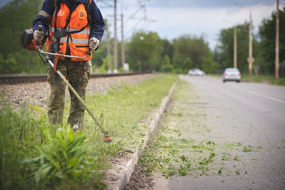 A man is using a lawn mower to cut grass on the side of the road.