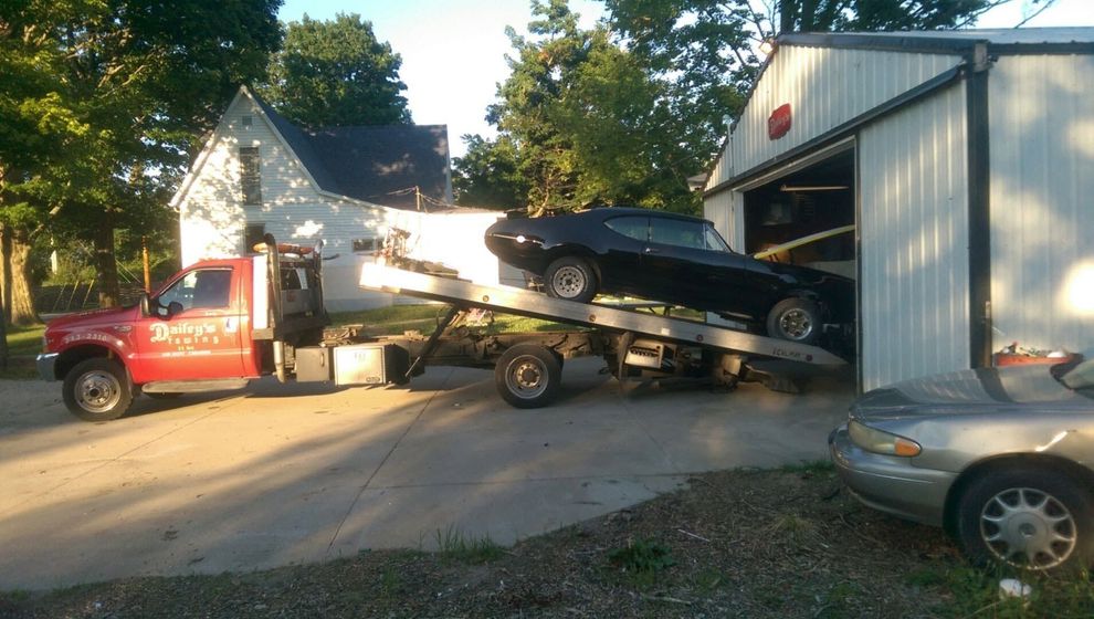 Red car being towed on a flatbed tow truck on a road; hazard triangle in front.