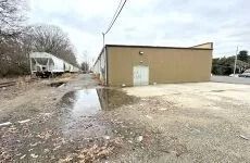 Empty gravel lot beside a tan building, with standing water and a white trailer in the background.