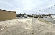 Empty gravel lot beside a tan building, with utility poles, parked vehicles, and an overcast sky.