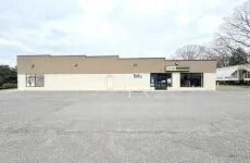 Small beige commercial building with a parking lot in front, viewed from a distance.