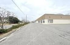 Quiet street beside a beige warehouse on a cloudy day, with bare trees and power lines.