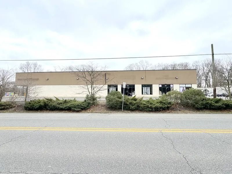 Low tan building behind shrubs across a paved road under a cloudy sky