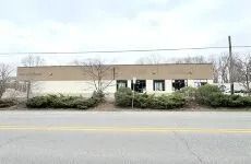 Single-story beige building across a road, with windows, shrubs, and bare trees under an overcast sky