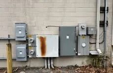 Wall-mounted utility boxes and electrical meters on a beige exterior brick wall, with visible rust stains.