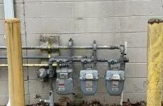 Gas meters and pipes mounted on a cinderblock wall behind yellow bollards.