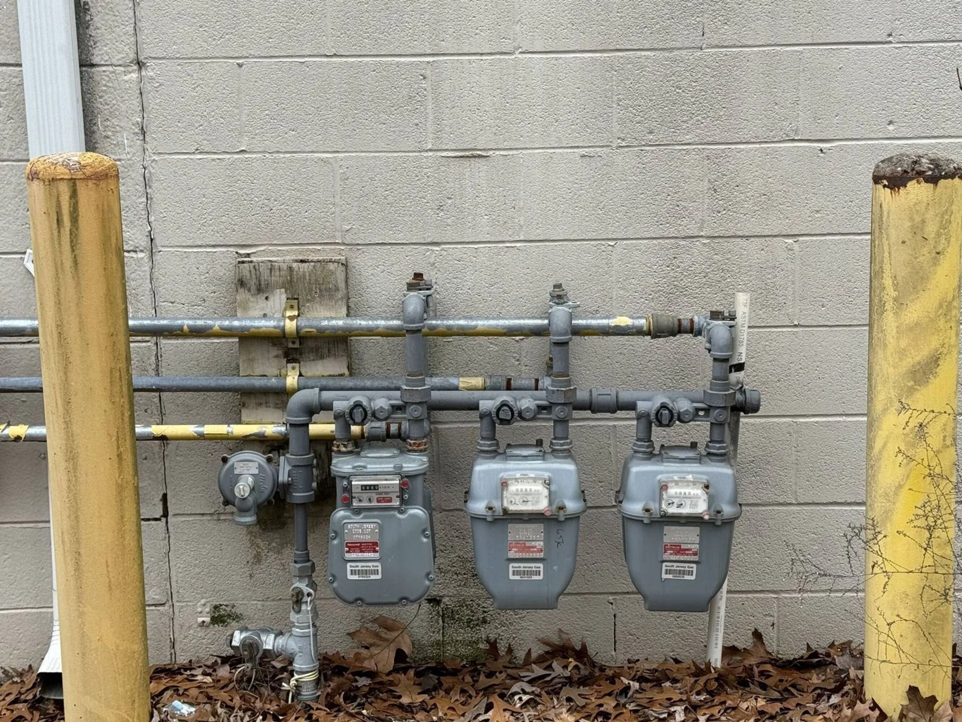Outdoor gas meter bank with three gray meters and yellow safety posts against a concrete wall