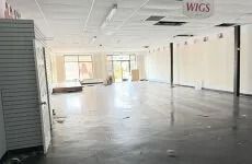 Empty retail store interior with tiled floor, large windows, and a “WIGS” sign above the wall