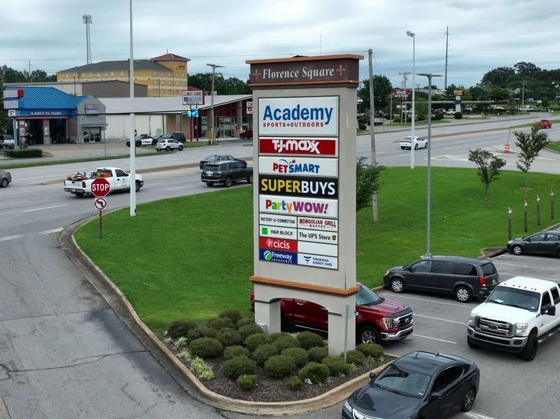 Shopping center sign listing stores in a parking lot beside a road, with cars and green grass.