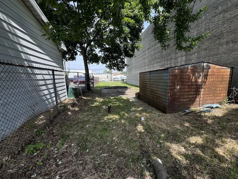 Narrow grassy alley between two buildings, with a brown shed and trees overhead.