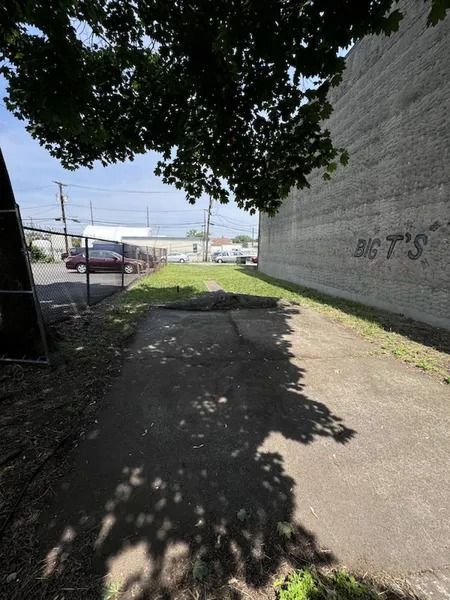 Shaded sidewalk beside a brick wall, opening to a sunny street and parking area.