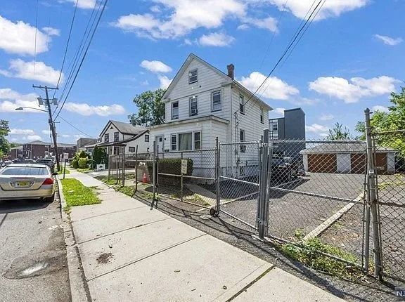 White two-story house behind chain-link fence beside sidewalk and parked cars on a sunny street.