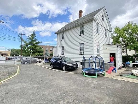 White house beside a driveway with a black car, trees, and a small blue trampoline in the yard