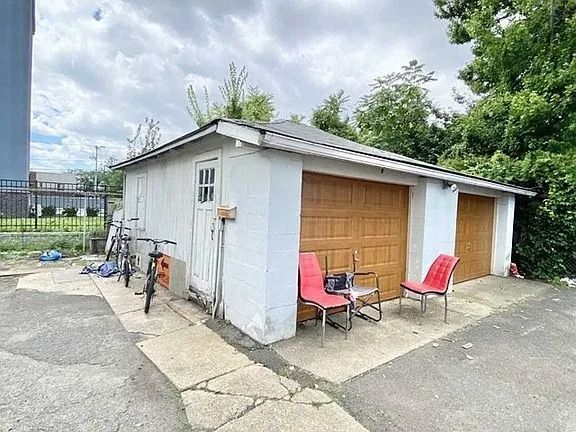 Small garage with white siding and tan door, two red chairs outside on a cracked driveway.