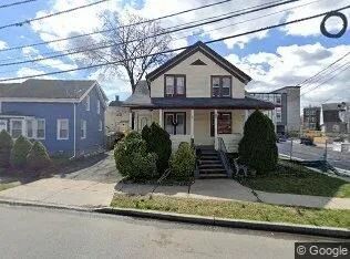 Small two-story beige house with front porch, shrubs, and a driveway on a residential street