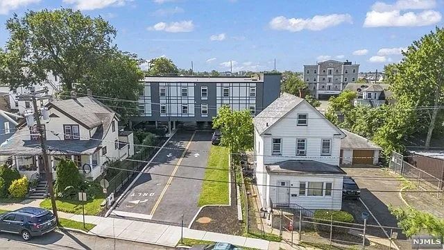 Aerial view of a residential street with houses, a central apartment building, and parked cars under a blue sky.