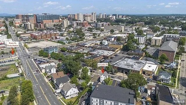 Aerial view of a city neighborhood with streets, buildings, trees, and a distant skyline under a blue sky