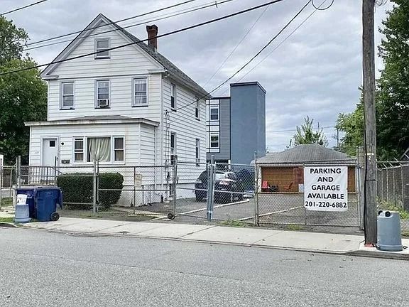 White house beside a fenced parking lot with a warning sign and blue trash bins on a street corner