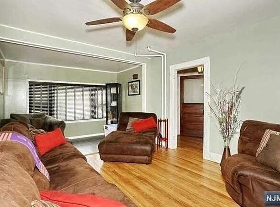 Bright living room with hardwood floors, brown chairs, red pillows, and a ceiling fan by a window and staircase.