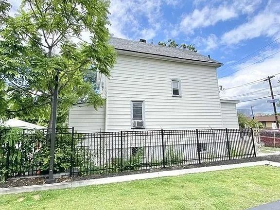 White house with black fence, green lawn, and tree-lined yard under a cloudy sky.