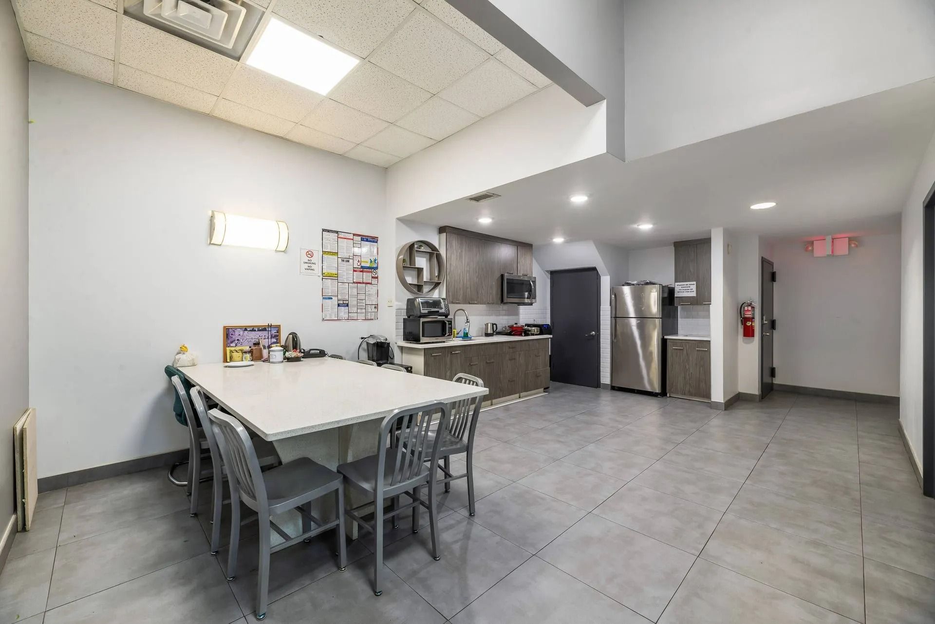 Bright break room with white tables, gray chairs, tiled floor, kitchenette, and refrigerator