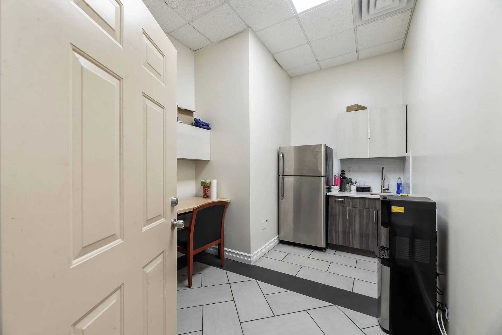Small office kitchenette with a stainless fridge, sink, cabinets, desk, and black water cooler.