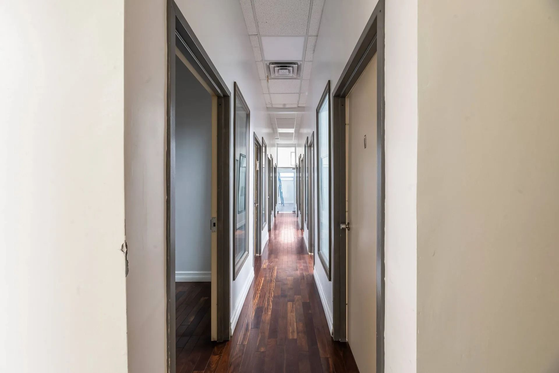 Narrow hallway with wooden floors and multiple doorways on both sides, lit by ceiling lights.