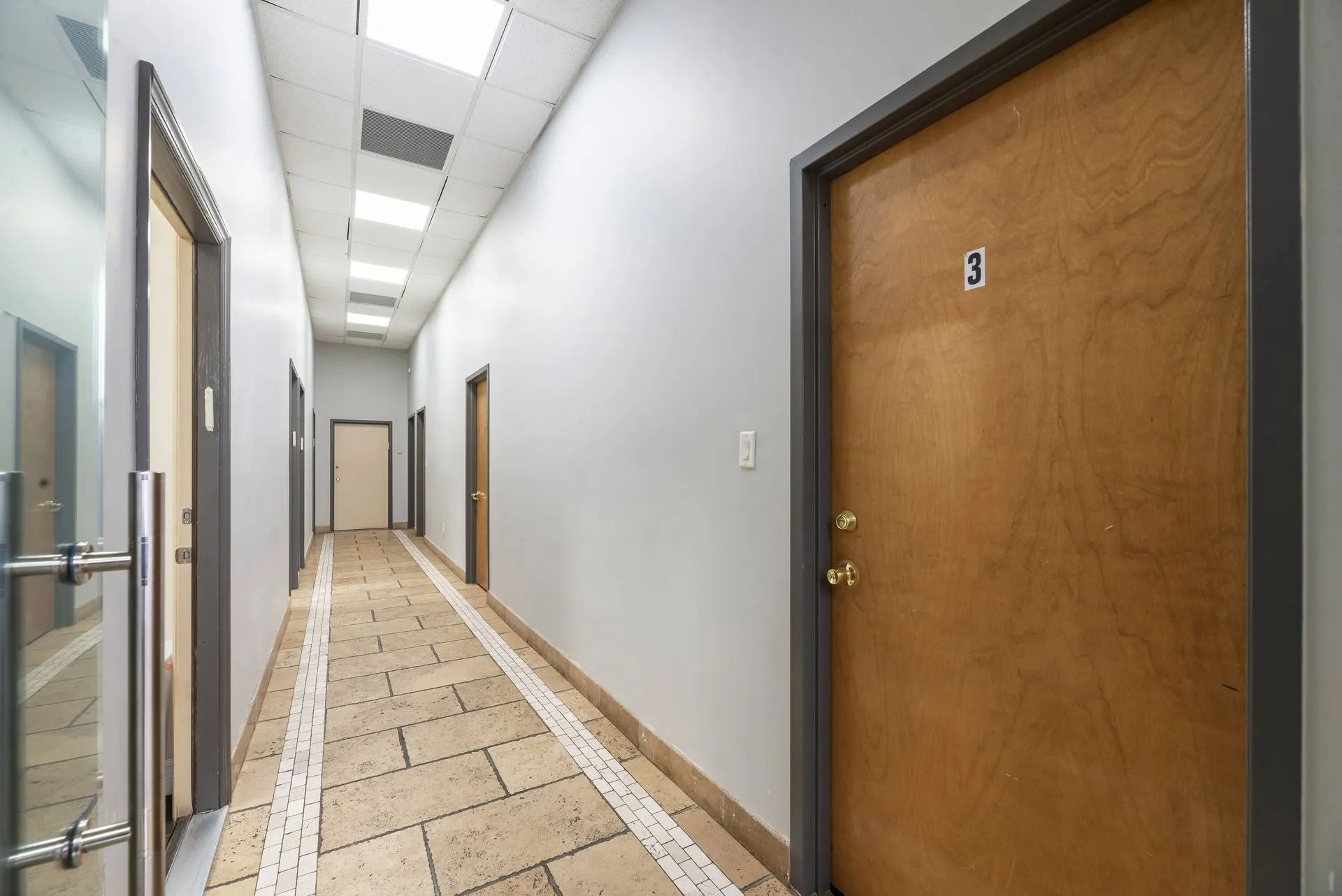 Empty office hallway with white walls, tan tile floor, and wood doors on both sides