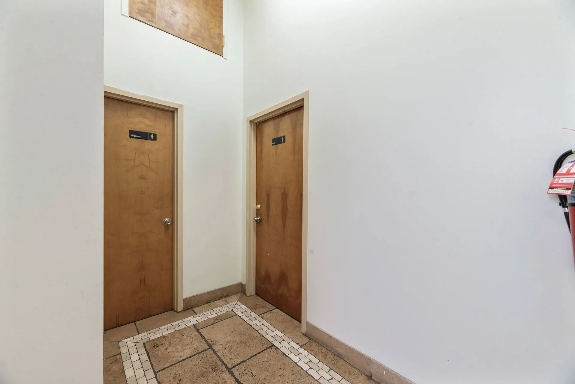 Narrow hallway with two wooden doors, white walls, and tiled floor under a bright skylight.
