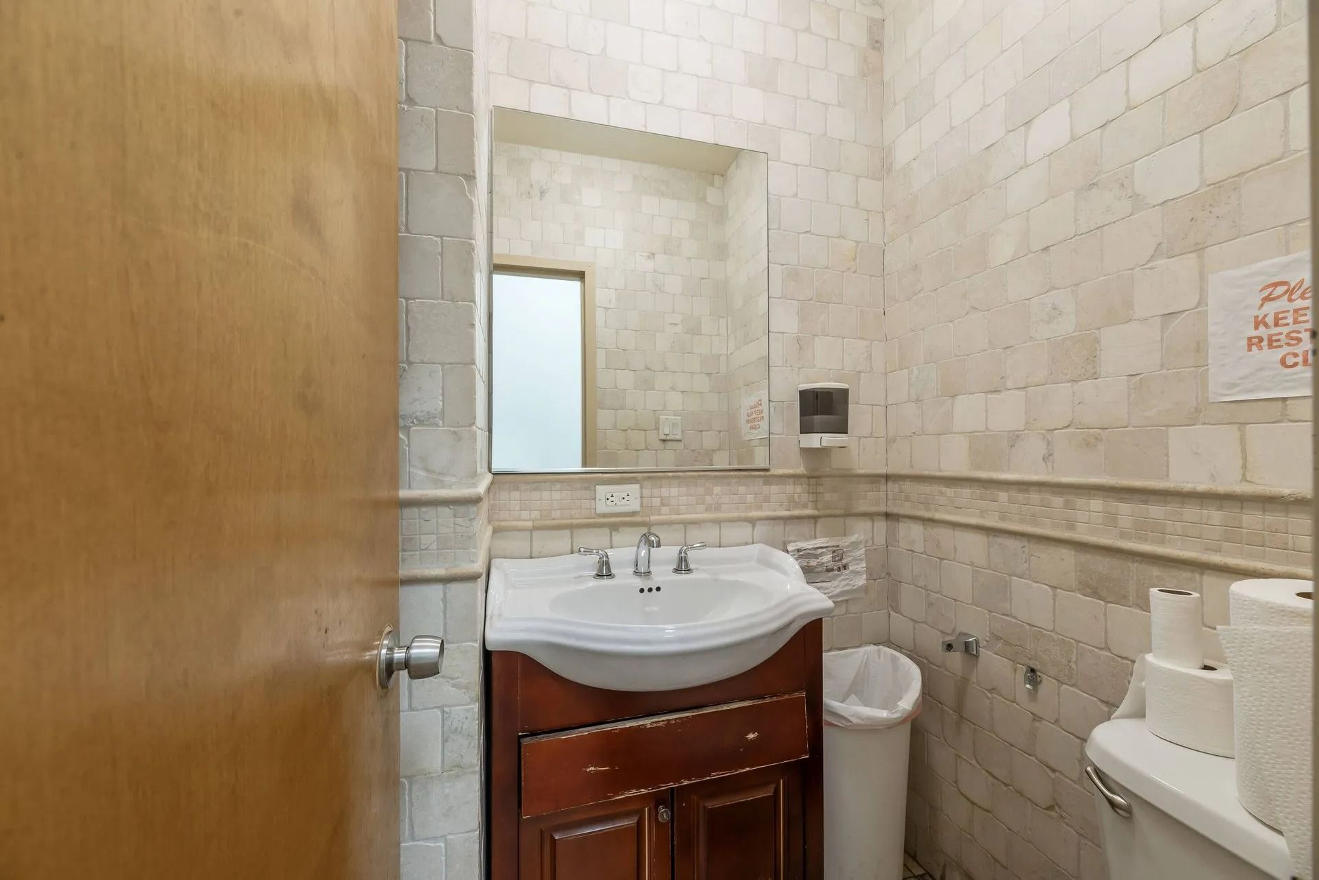 Small beige-tiled restroom with sink, mirror, toilet, and open wooden door