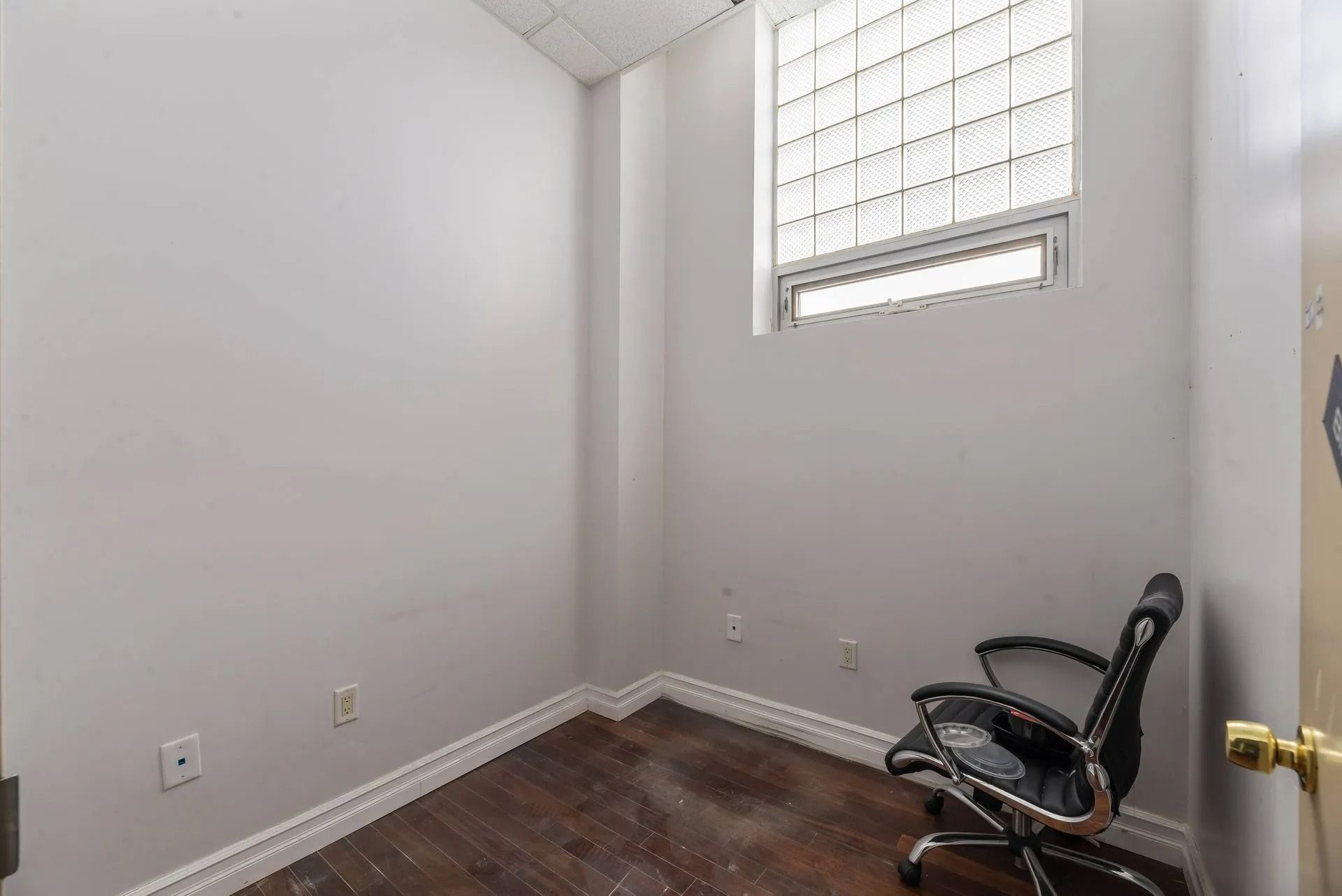 Small empty room with white walls, dark wood floor, and a black chair near a window.