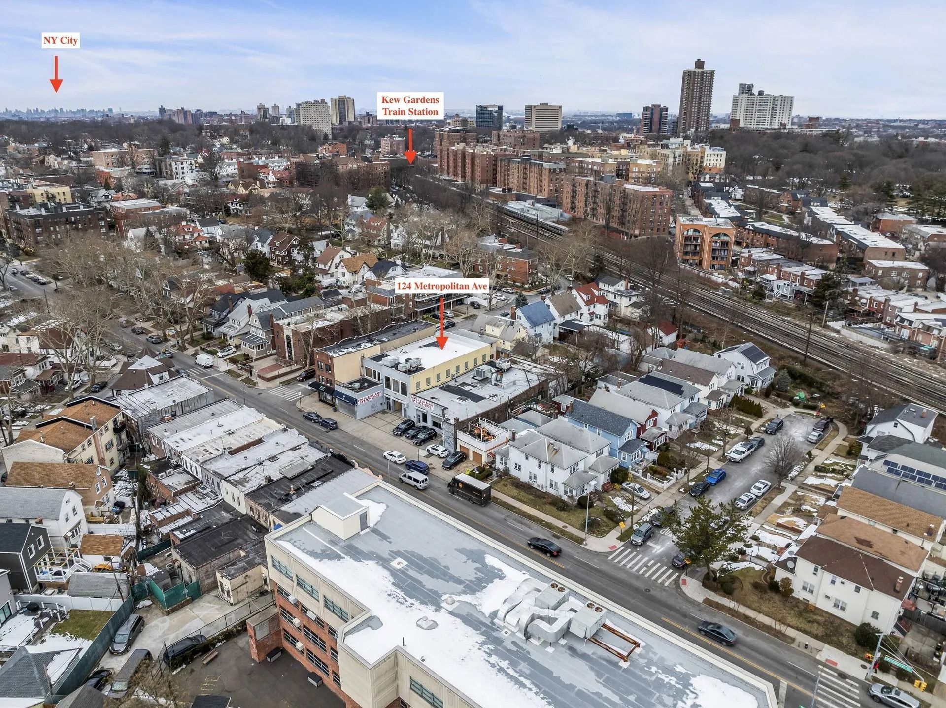 Aerial view of a snowy neighborhood with houses, roads, and distant city buildings under a cloudy sky