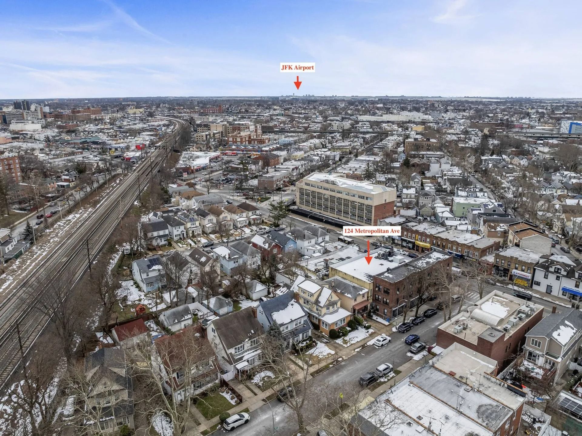 Snowy aerial view of a suburban neighborhood with houses, roads, and a horizon of buildings