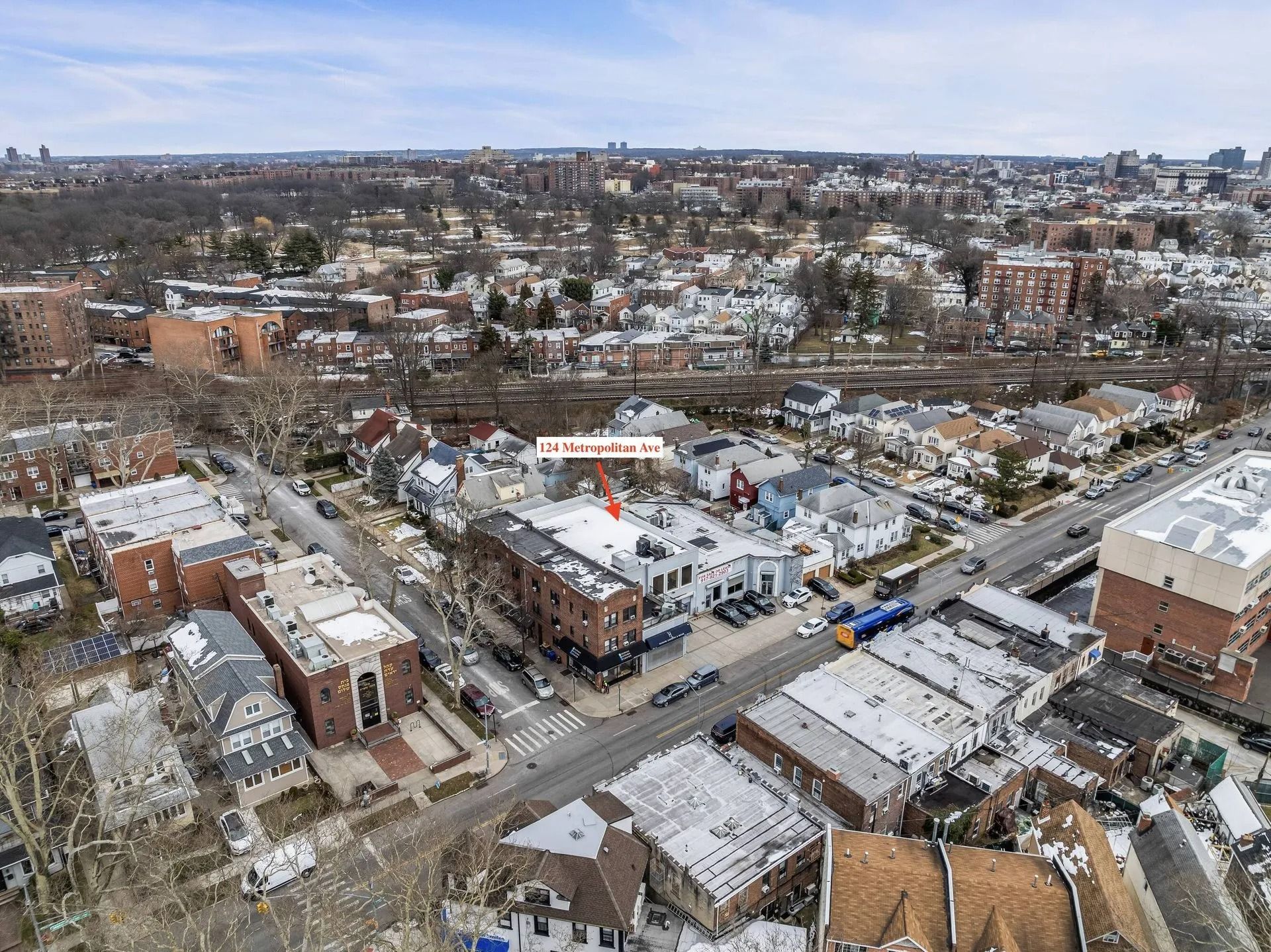Aerial view of a snowy urban neighborhood with streets, buildings, and a distant city skyline.