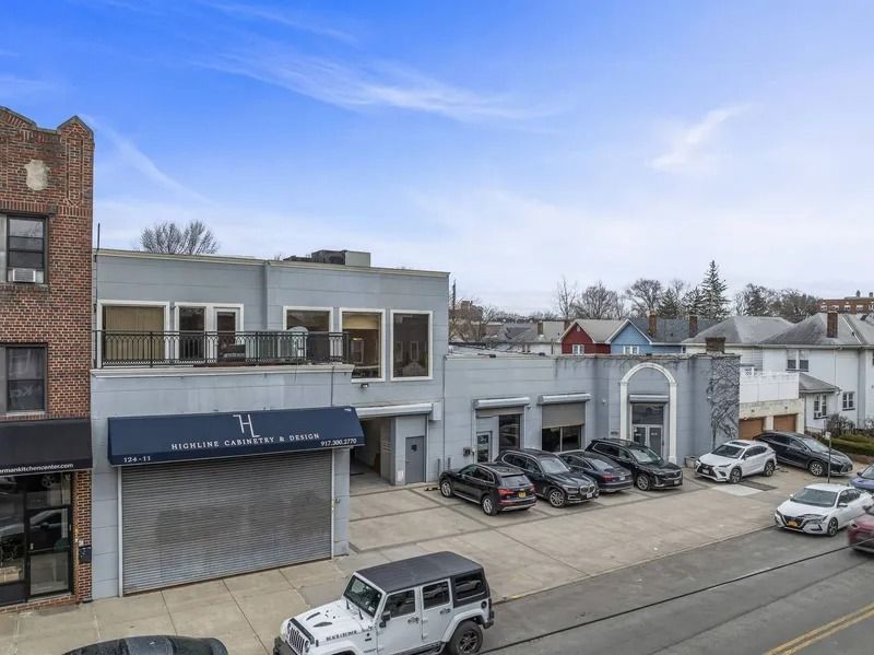 Street view of a gray commercial building with a blue awning, parked cars, and row houses behind it