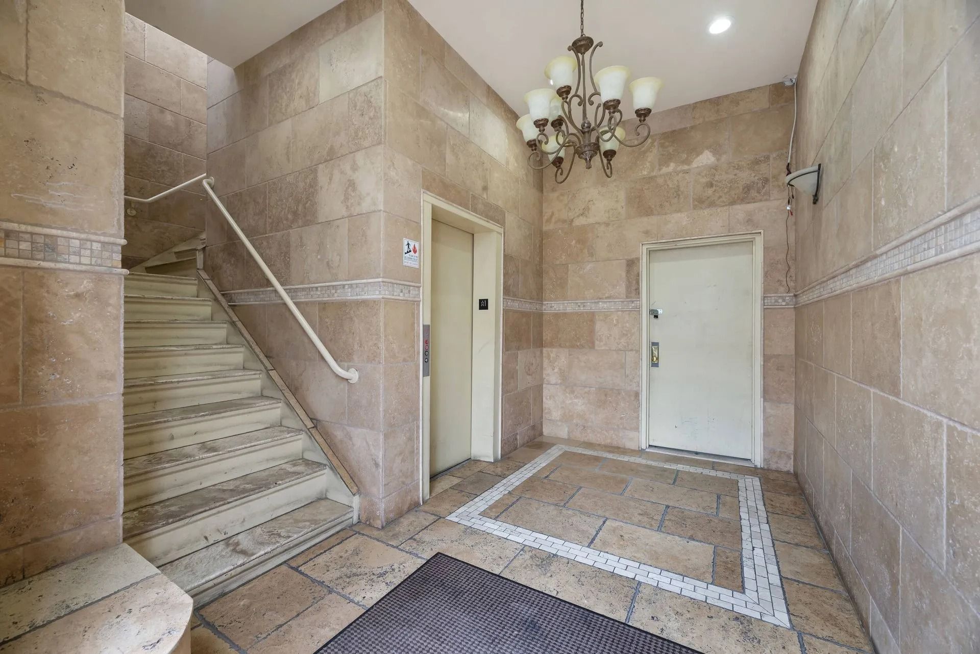 Stone apartment lobby with stairs, elevator doors, and a chandelier overhead