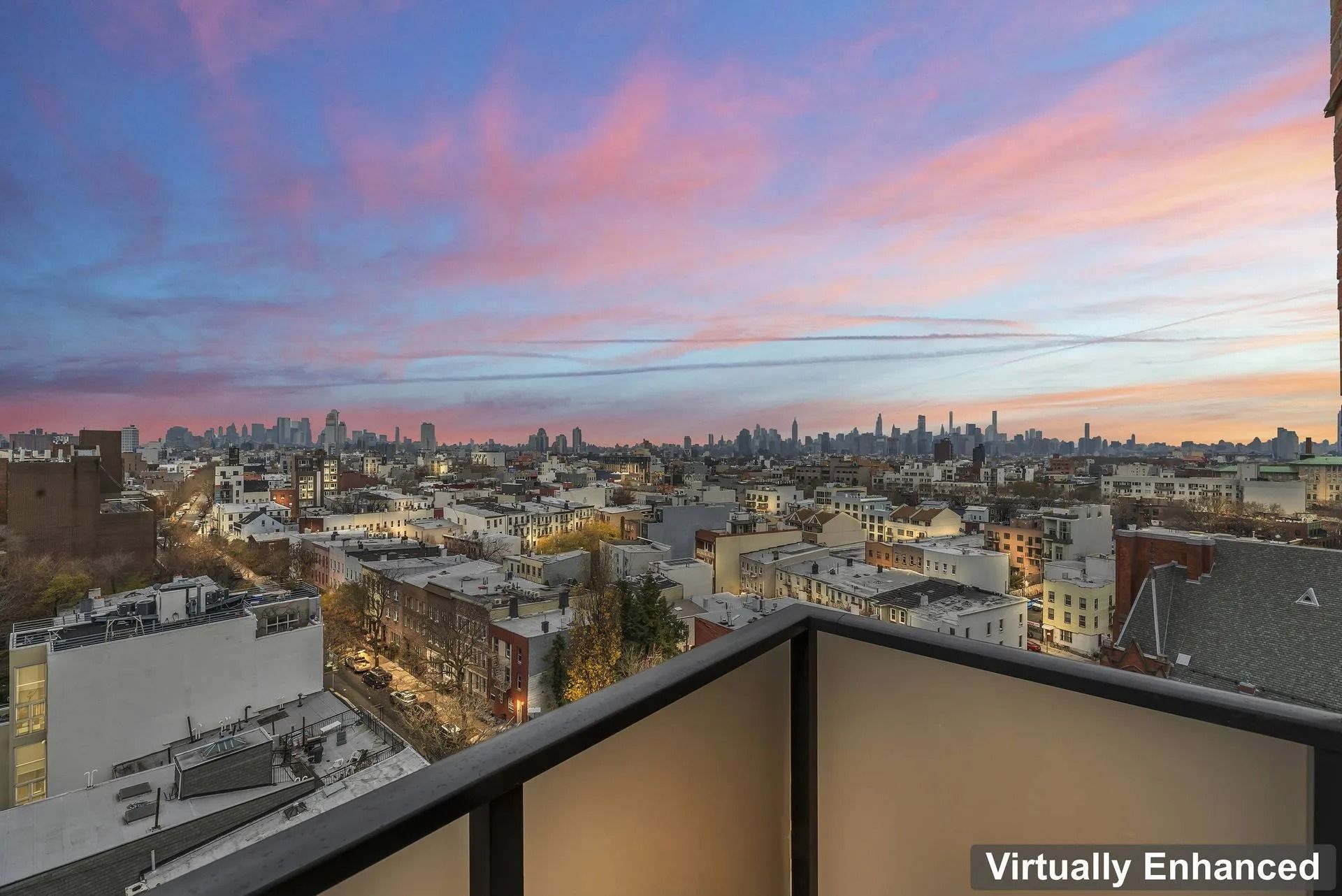 City skyline at sunset seen from a balcony, with pink and blue sky over rooftops and distant buildings