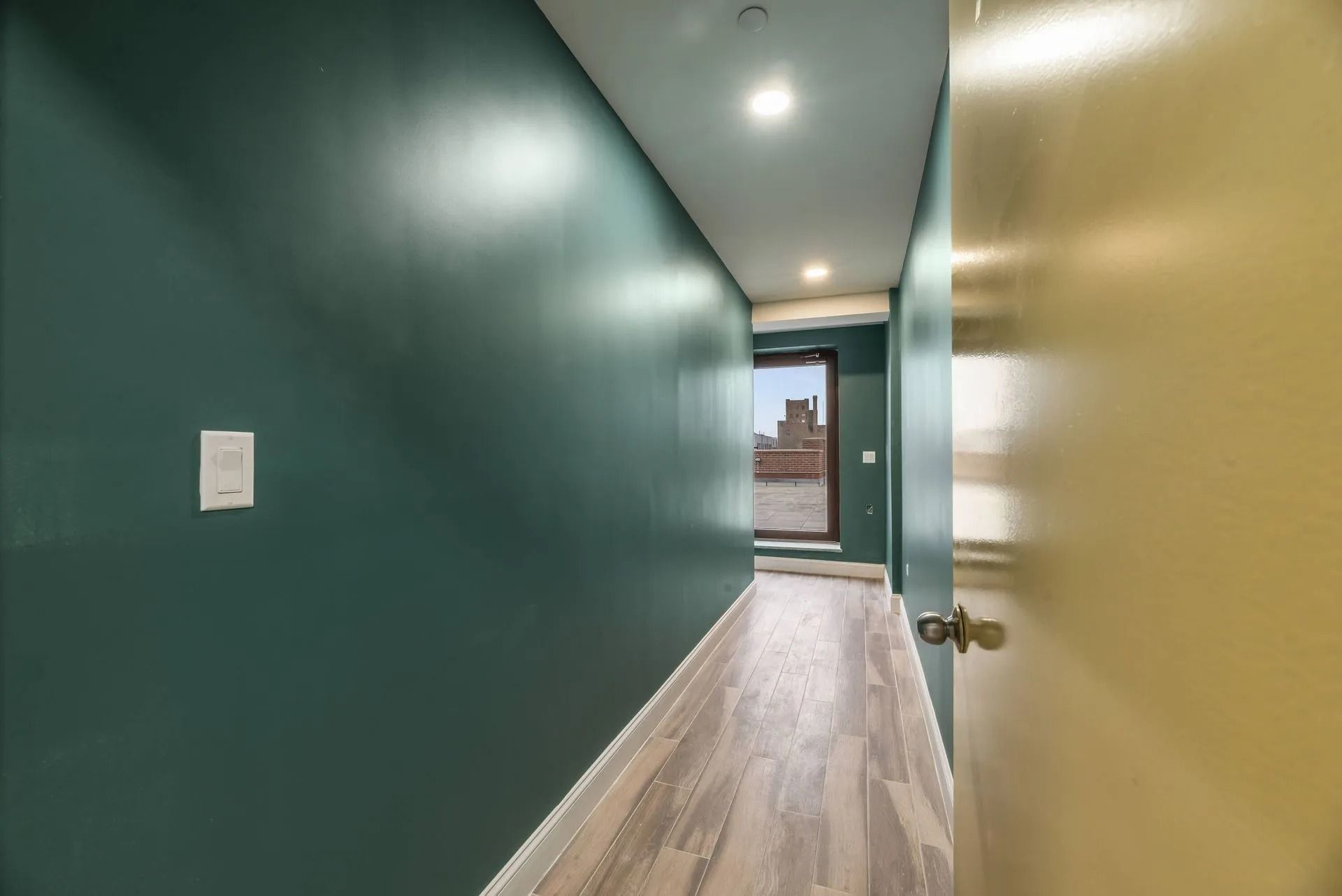 Narrow hallway with green walls, wood floor, and an open door leading to a bright room.