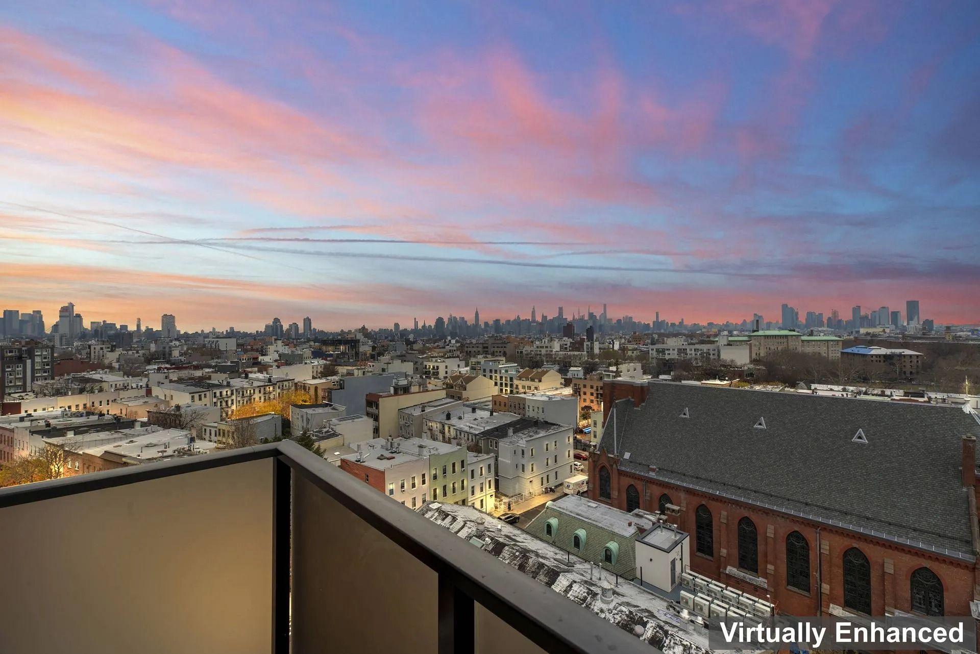 City skyline at sunset with pink clouds above rooftops and a balcony view in the foreground