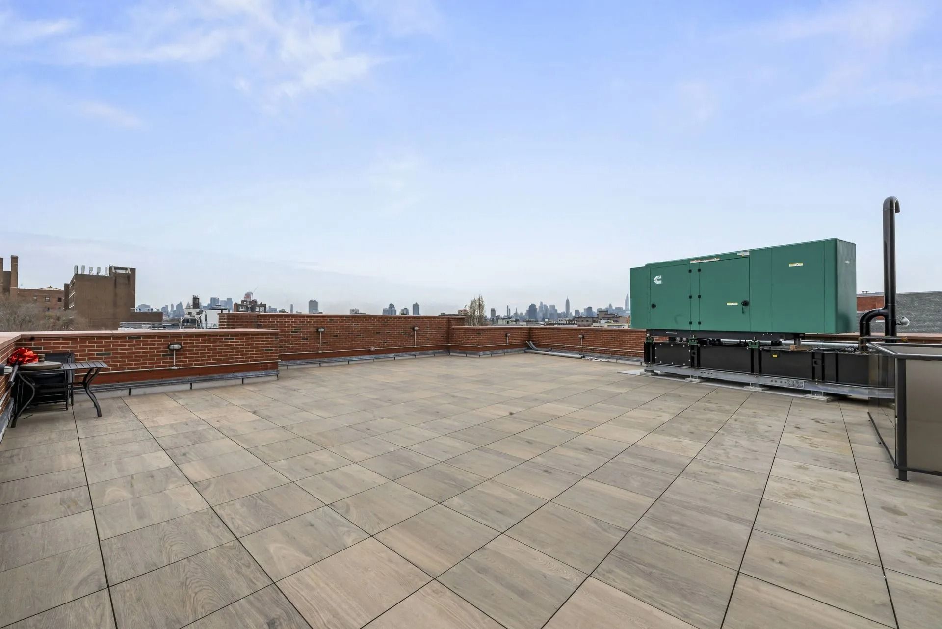 Rooftop terrace with gray pavers, brick wall, and green rooftop equipment under a cloudy sky