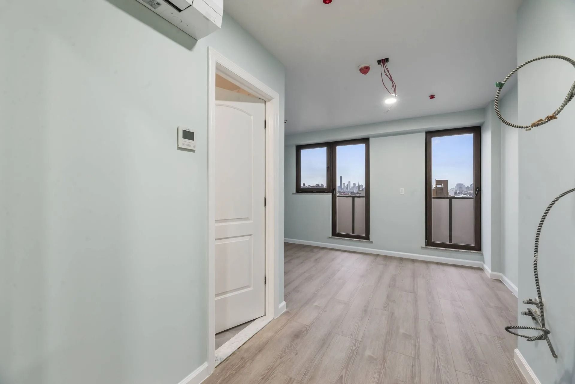 Bright empty room with white walls, light wood floors, and large windows.