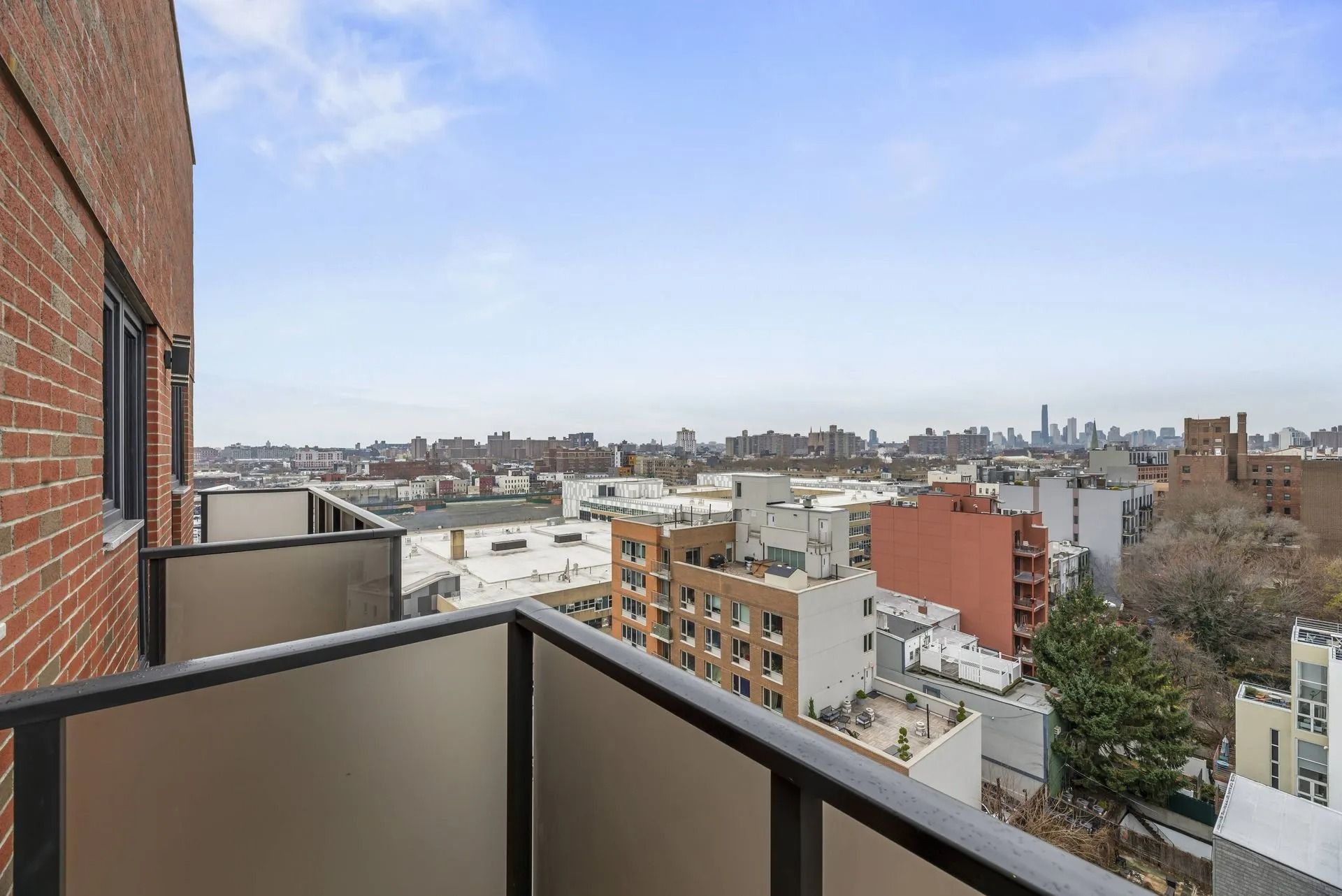 City skyline viewed from a balcony beside a brick building, with rooftops and a blue sky.
