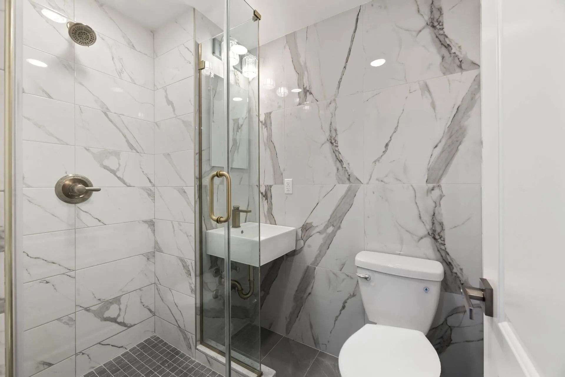 Compact modern bathroom with marble walls, glass shower, and white toilet next to a small sink.