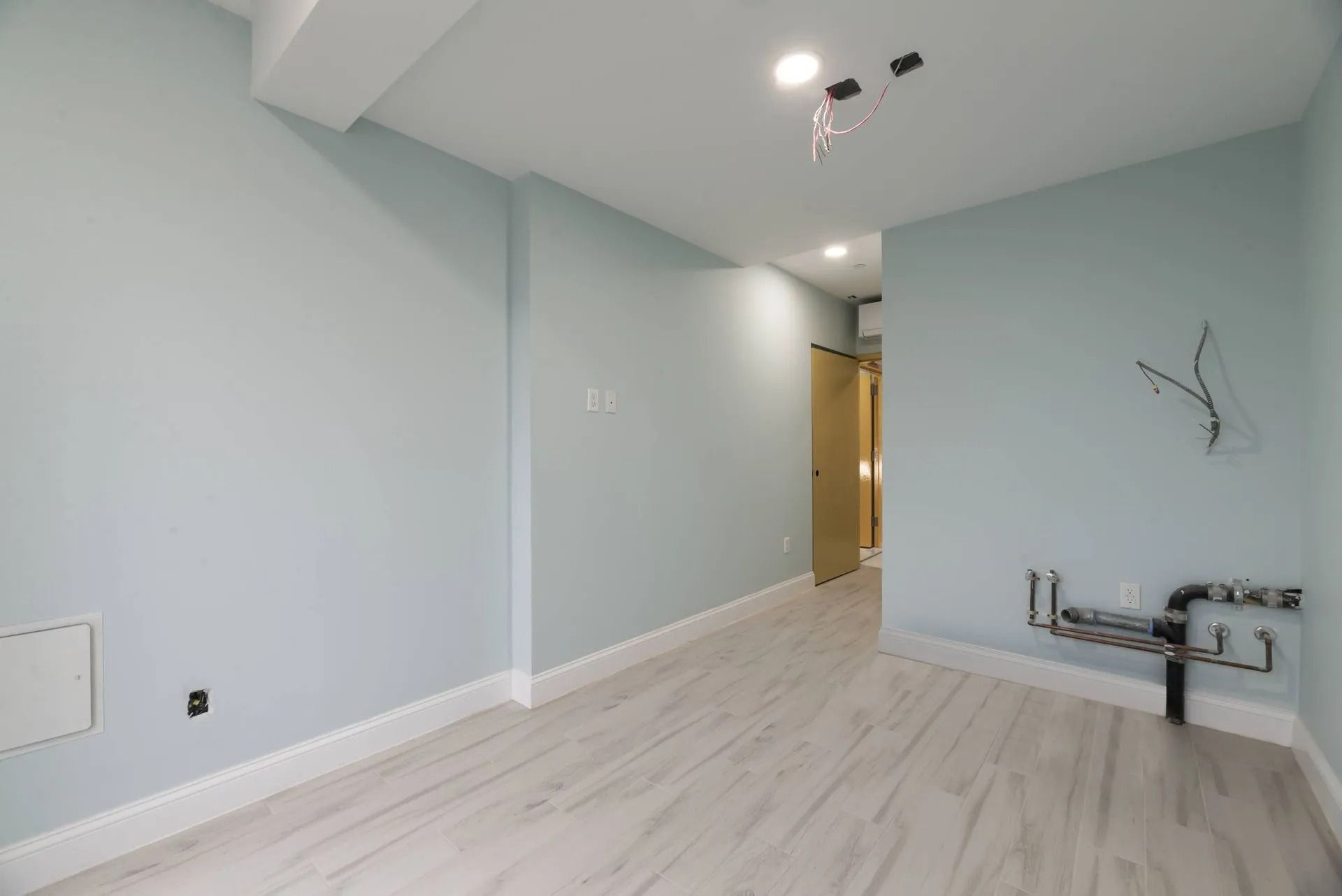 Empty light-gray room with white floors, ceiling lights, and exposed plumbing on the right wall