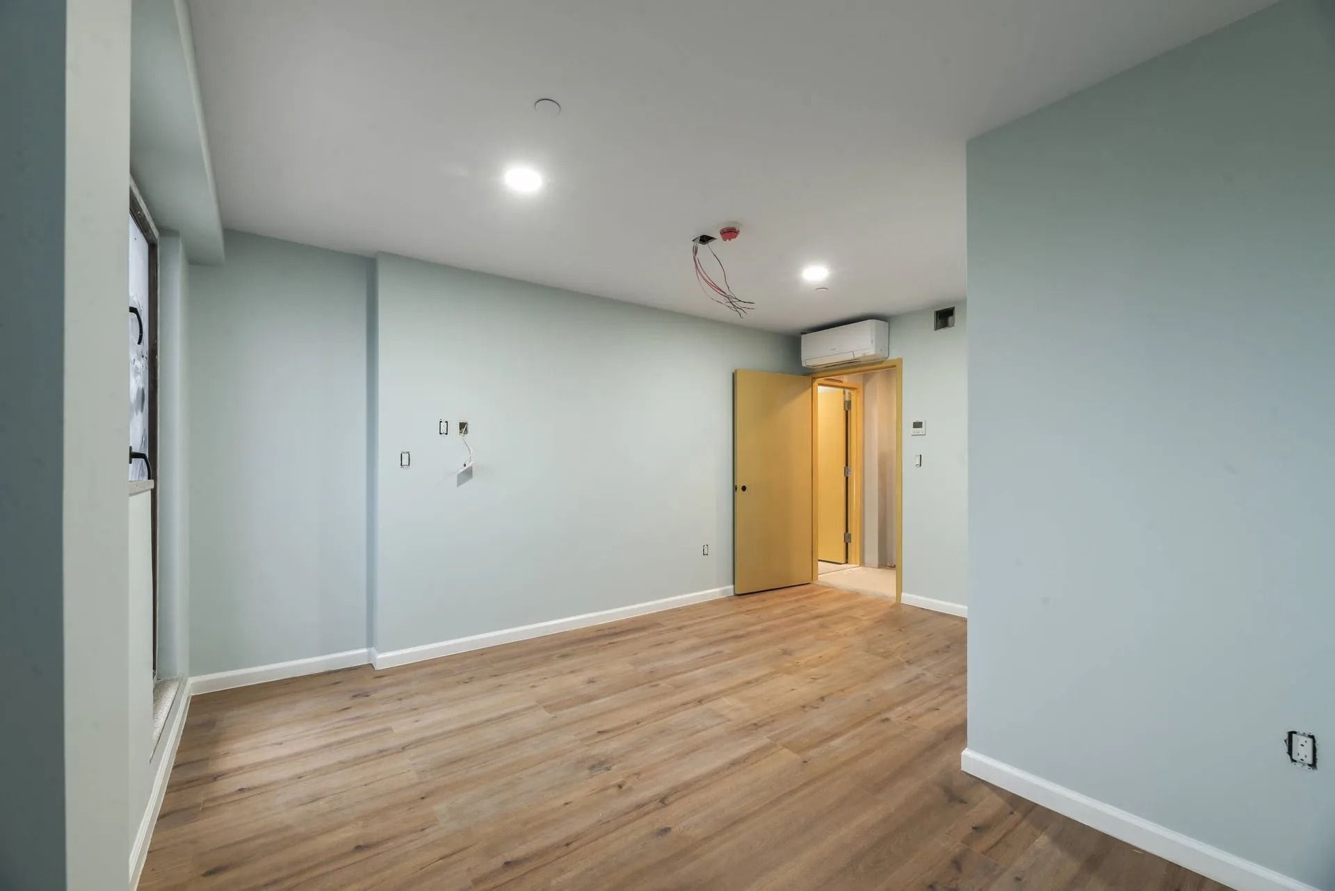 Empty modern room with light blue walls, wood floor, recessed lights, and a doorway at the back