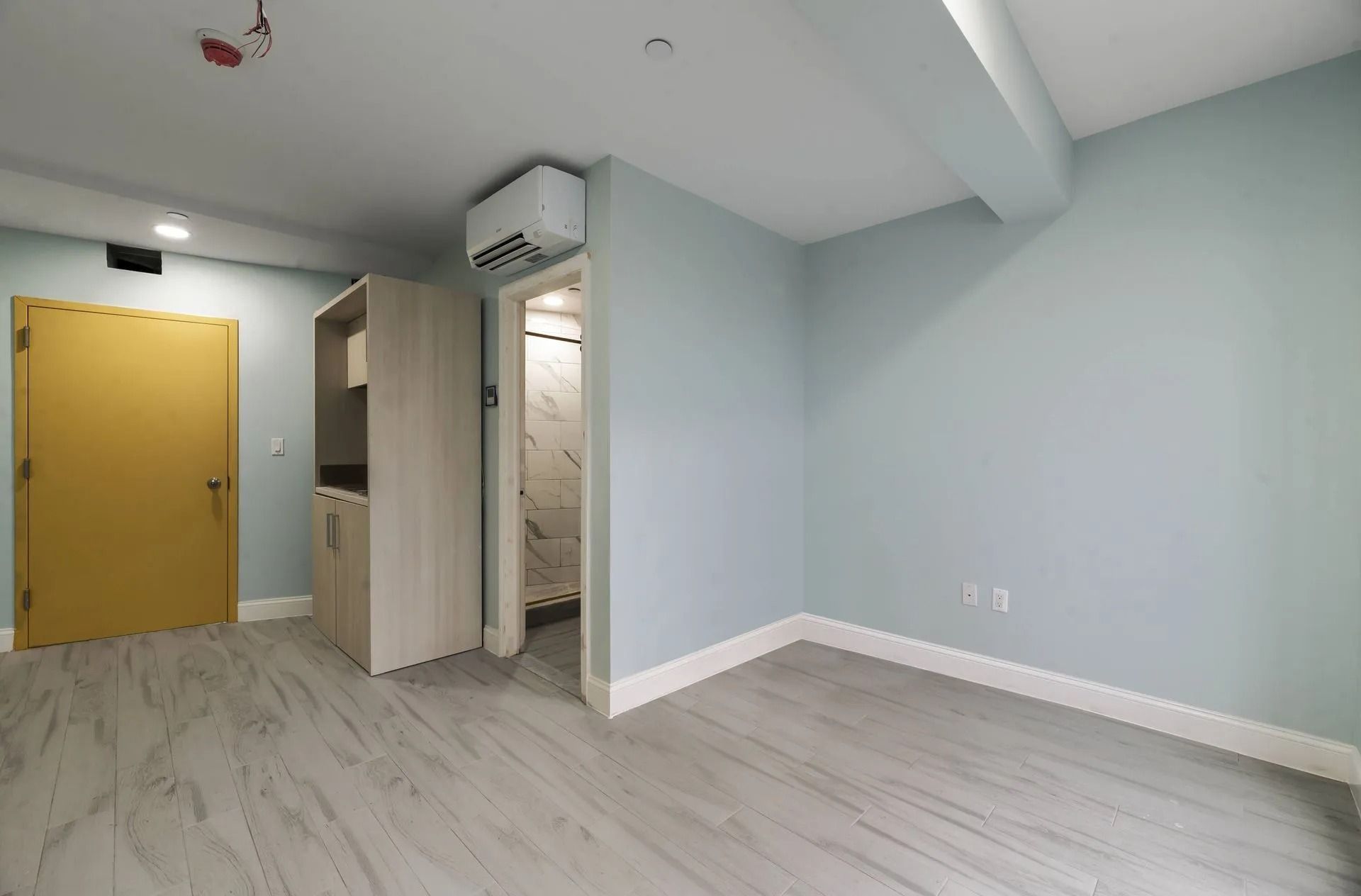 Empty modern room with pale blue walls, white wood floors, yellow door, and open bathroom doorway