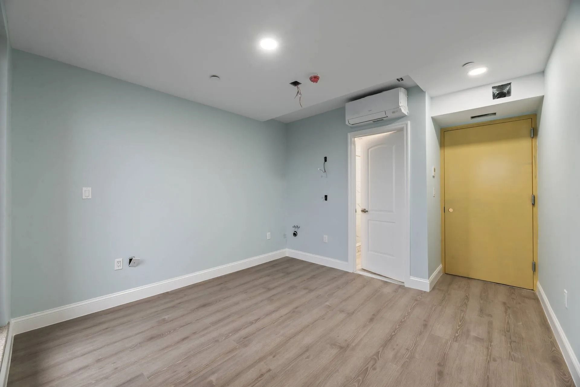 Empty light-blue room with wood floor, recessed lights, and a yellow door