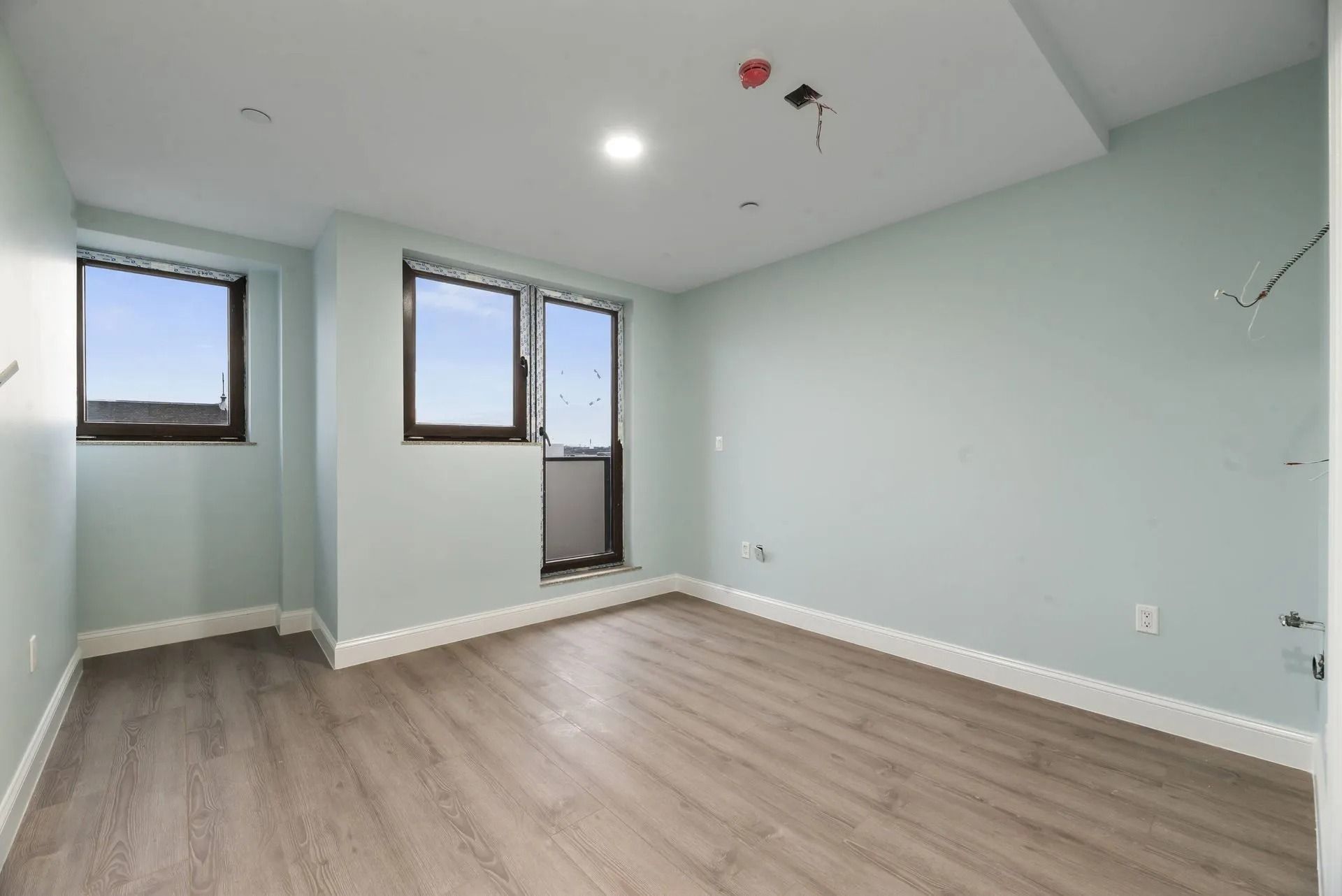 Empty light-blue room with wood flooring, two windows, and a glass door.