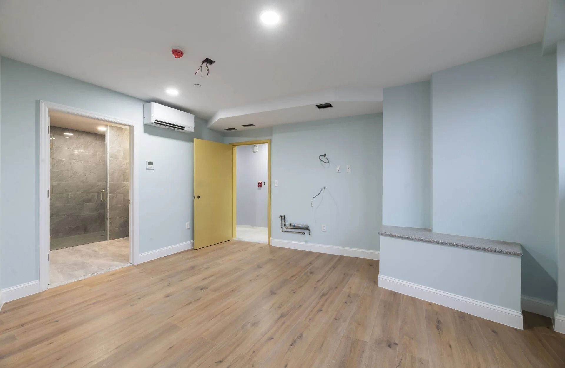 Empty light-blue room with wood floors, white ceiling, yellow door, and a tiled bathroom area on the left.