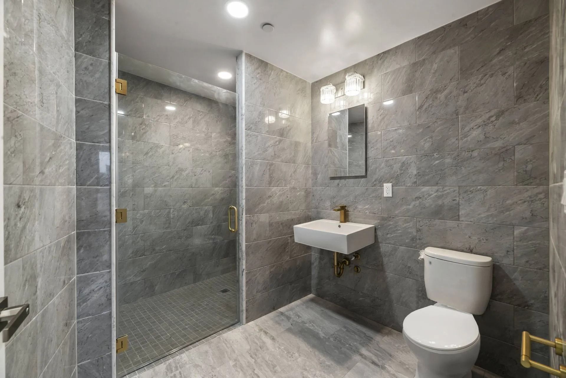 Modern gray-tiled bathroom with glass shower, wall sink, mirror, and toilet